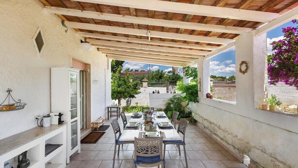 A covered patio with a dining table set for four, surrounded by chairs. A white cabinet and a small shelf with a basket are visible on the left.