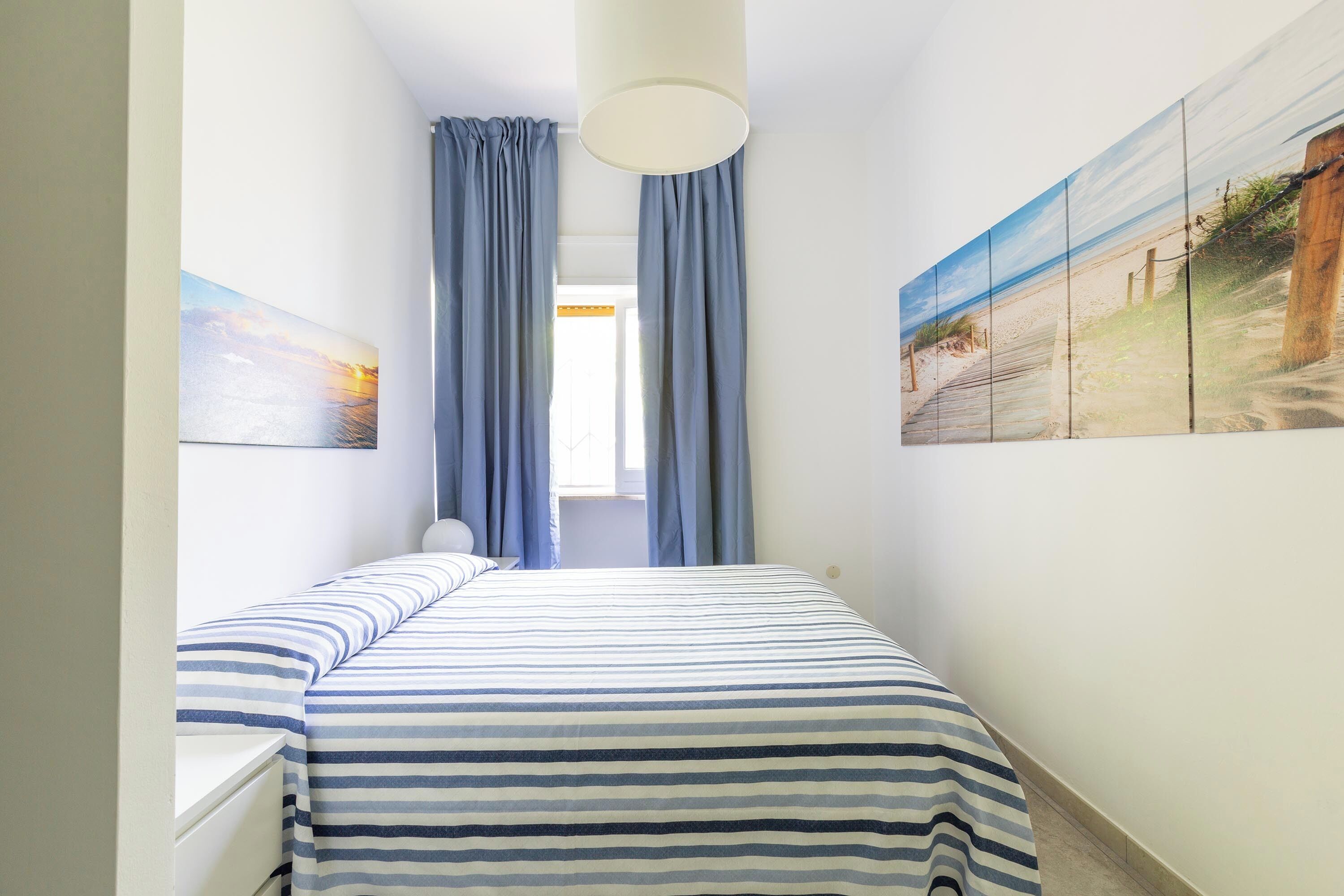 A narrow hotel room with a bed, a nightstand, a lamp, and a large painting on the wall.