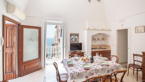 Uma área de refeições com uma mesa posta para quatro pessoas, cozinha ao fundo e vista para o mar por uma porta aberta.
