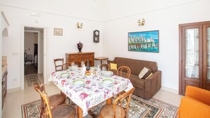 Uma área de refeições com uma mesa posta para quatro pessoas, rodeada por cadeiras de madeira. Um sofá castanho e um poltrona laranja estão posicionados junto à parede.