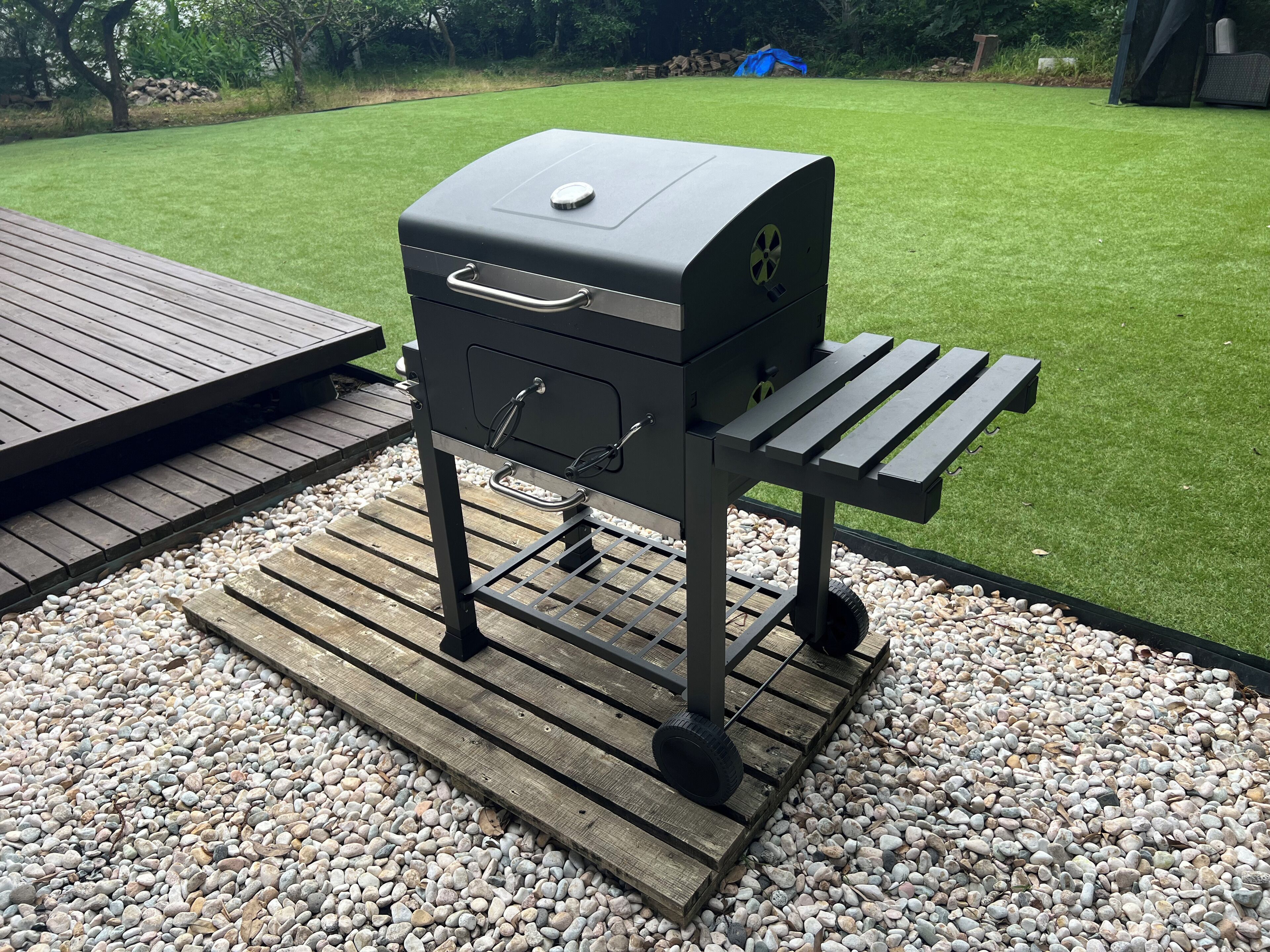 A barbecue grill with a metal frame, a lid, and a side shelf, placed on a wooden deck with a gravel base.
