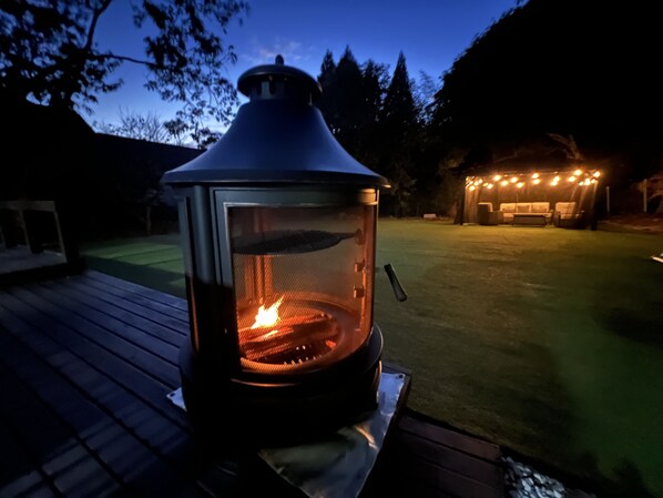 A wood-burning stove on a deck with a fire inside, surrounded by a deck and a grassy yard.