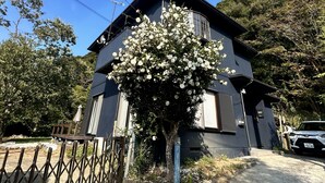Un edificio de dos pisos con fachada oscura, un balcón con barandilla de madera y un árbol con flores blancas en primer plano.
