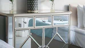 Ein Nachttisch mit Spiegel, mit einer zentralen Dekoflasche, flankiert von zwei Vasen mit weißen Blumen, der einen Blick ins Schlafzimmer reflektiert.