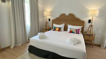 A neatly made bed with white linens and colorful pillows, two bedside tables with lamps, a large window with curtains, and a wooden ceiling.