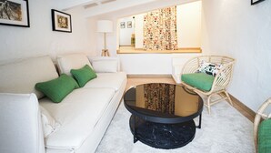 A modern living room with a white sofa, green cushions, a round glass coffee table, wicker chairs, and a large window with patterned curtains. - Hotel Maia Menorca (Ciutadella de Menorca)