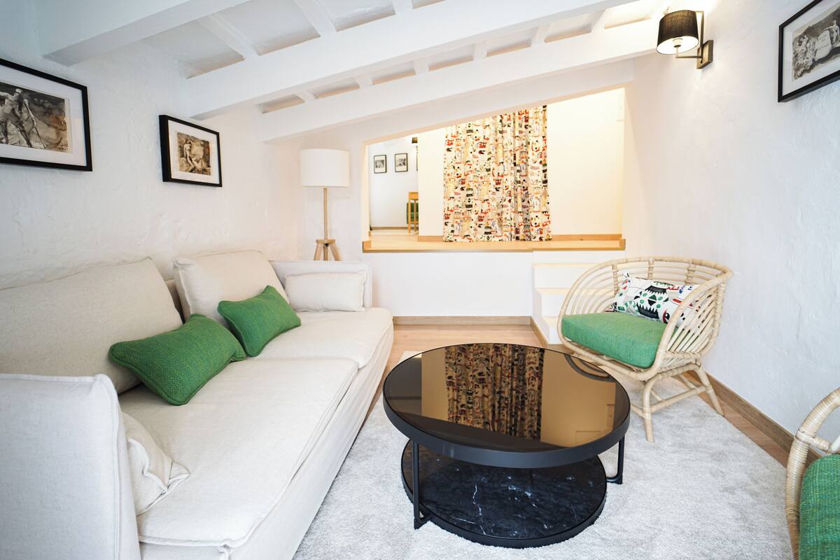 Una sala de estar moderna con un sofá blanco, cojines verdes, una mesa de centro redonda de vidrio, sillas de mimbre y un ventanal con cortinas estampadas.