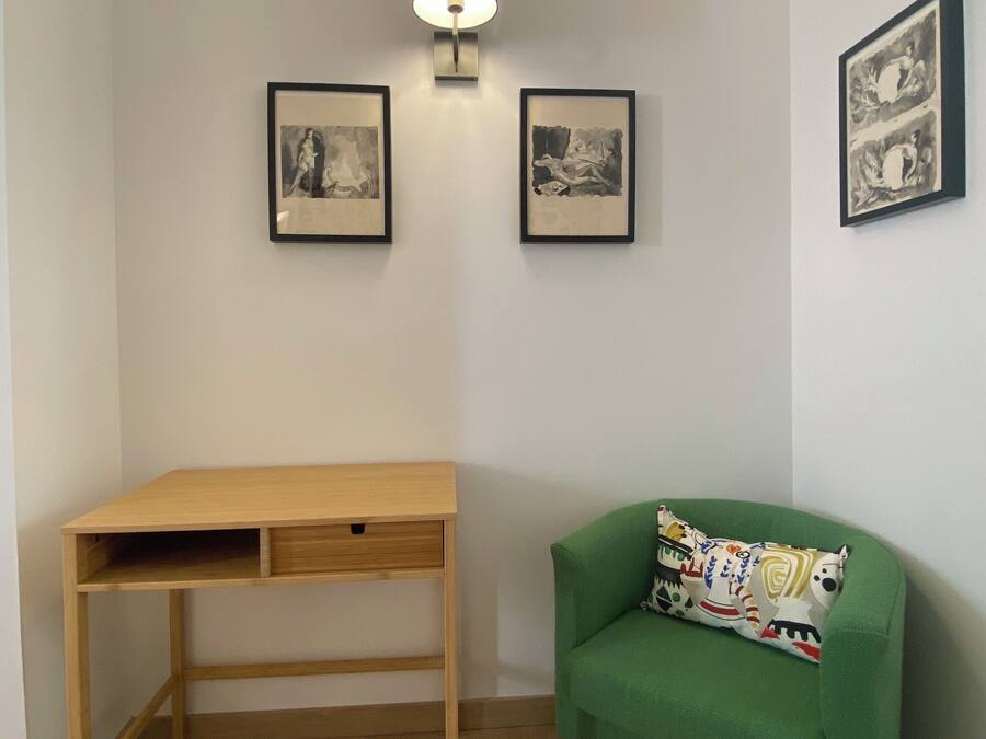 Una habitación con un escritorio de madera, un sofá verde con un cojín estampado y tres obras de arte enmarcadas en la pared.