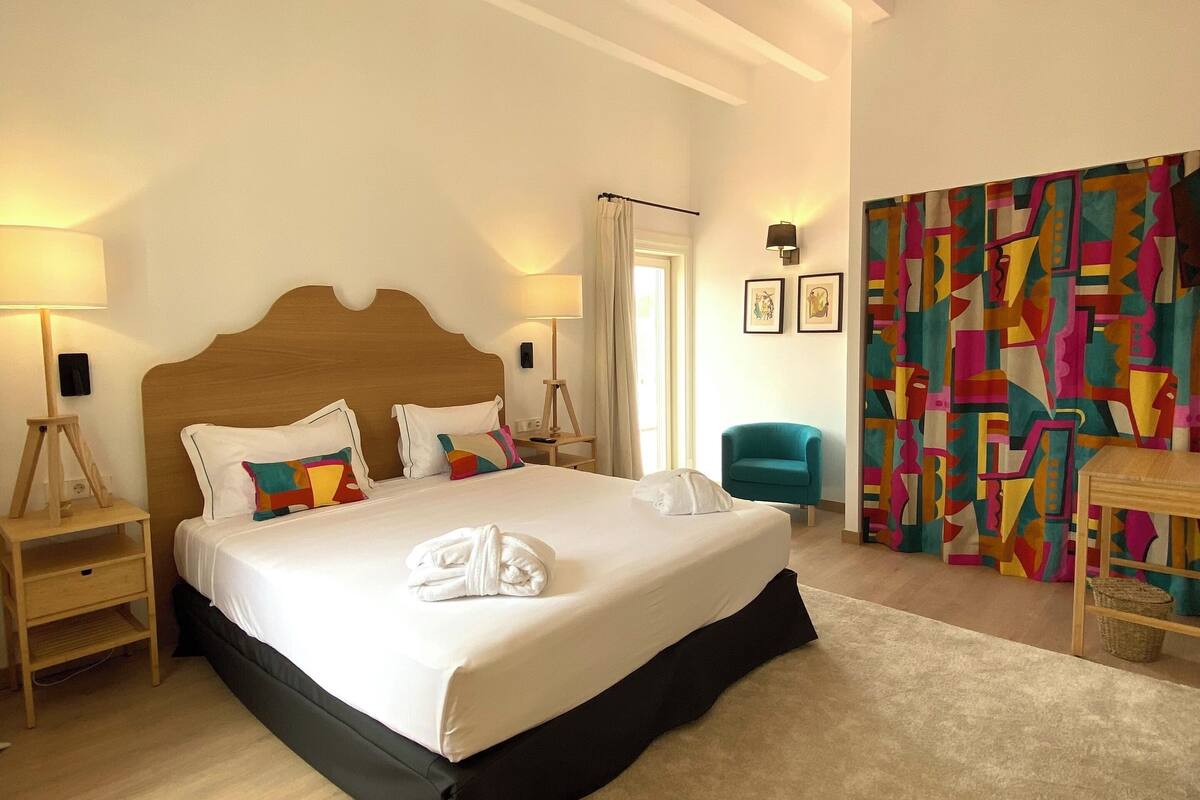 Un dormitorio con una cama grande, cabecera de madera, mesita de noche con lámpara, una silla y un colorido tapiz abstracto en la pared.
