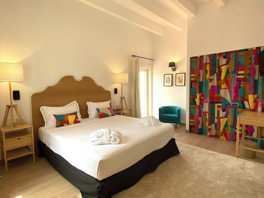 A bedroom with a large bed, a wooden headboard, a bedside table with a lamp, a chair, and a colorful abstract wall hanging.