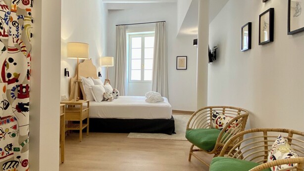 A hotel room with a bed, a nightstand, a lamp, wicker chairs, and a view of the hallway. - Hotel Maia Menorca (Ciutadella de Menorca)