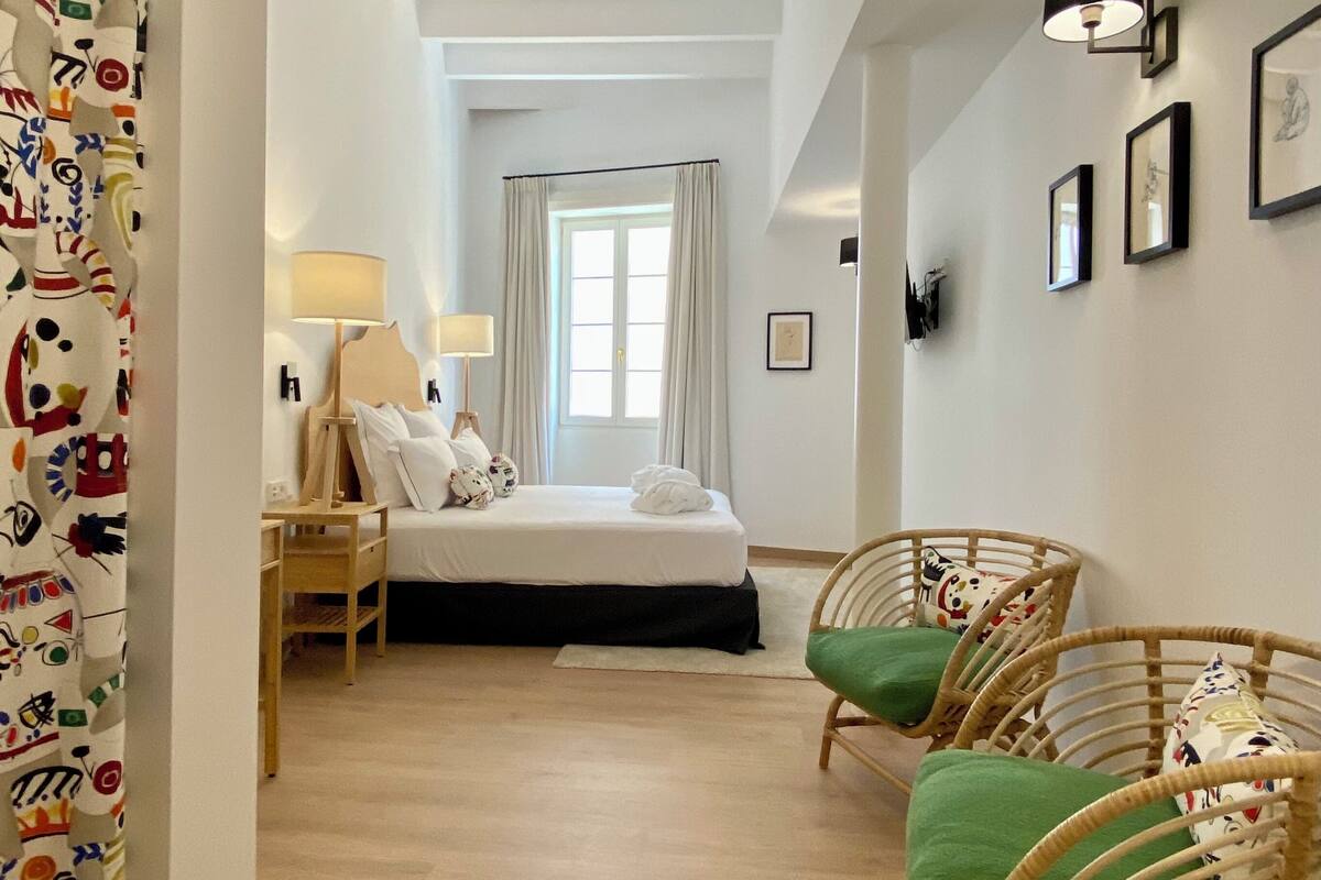 Habitación de hotel con cama, mesita de noche, lámpara, sillas de mimbre y vista al pasillo.