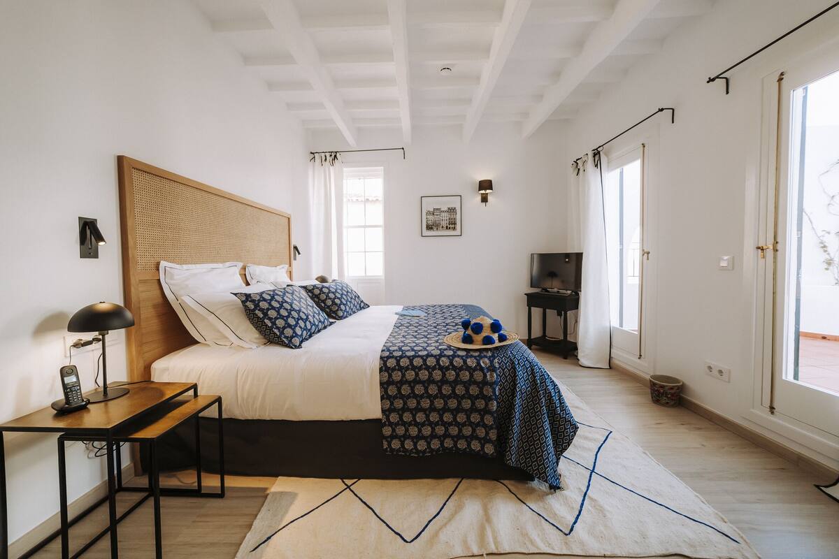 A bedroom with a bed, bedside table, lamp, and a view of the outdoors.