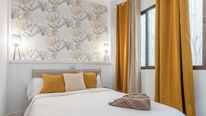 A neatly made bed with white and mustard bedding, a patterned wallpaper, and a window with yellow curtains.