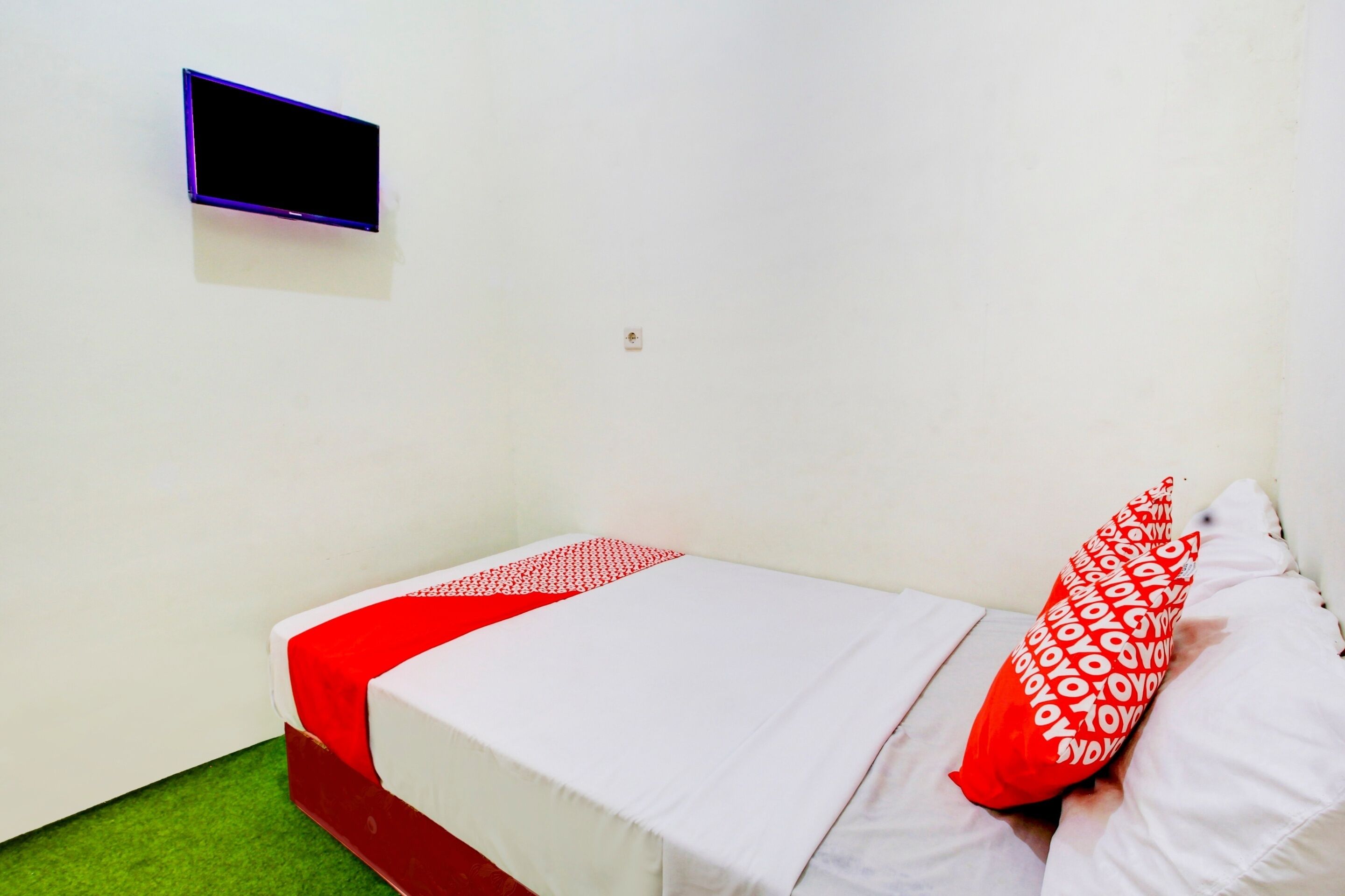 A single-bed room with a red and white bedspread, a red pillow with white text, and a small TV mounted on the wall.