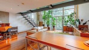 A modern interior with a large wooden table, a staircase, and large windows with plants. - Incredible 2BD Loft by Regents Canal - Haggerston (London)