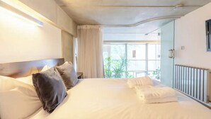A hotel room with a large bed, a headboard, a bedside table, a window with curtains, and a view of greenery outside. - Incredible 2BD Loft by Regents Canal - Haggerston (London)