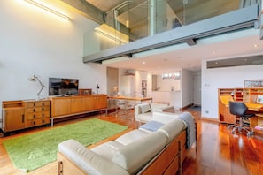 A spacious living area with a high ceiling, a green rug, a sofa, a TV, a wooden cabinet, and a kitchen in the background.