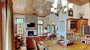 A spacious living area with a stone fireplace, wooden ceiling, and a dining table with chairs. - At the Start of It All by Taylor Made Deep Creek (McHenry)