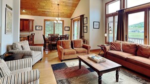 A spacious living room with a high ceiling, wooden beams, and large windows offering a view of the outdoors. - At the Start of It All by Taylor Made Deep Creek (McHenry)