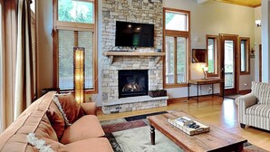 A spacious living room with a stone fireplace, a large flat-screen TV mounted above it, and a cozy seating area with a sofa and armchairs. - At the Start of It All by Taylor Made Deep Creek (McHenry)
