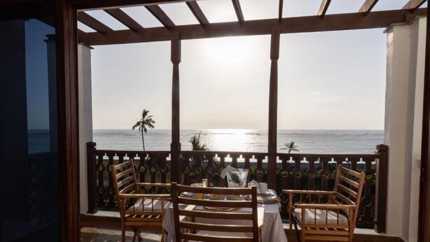 A balcony with wooden chairs and a table, overlooking a calm sea and palm trees. - The Sultan Palace Beach Retreat Three-Bedroom Villa (Kikambala, Mombasa)