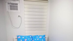 Un lit avec une literie bleue et des coussins à motifs floraux, une serviette enroulée et une fenêtre avec des stores.