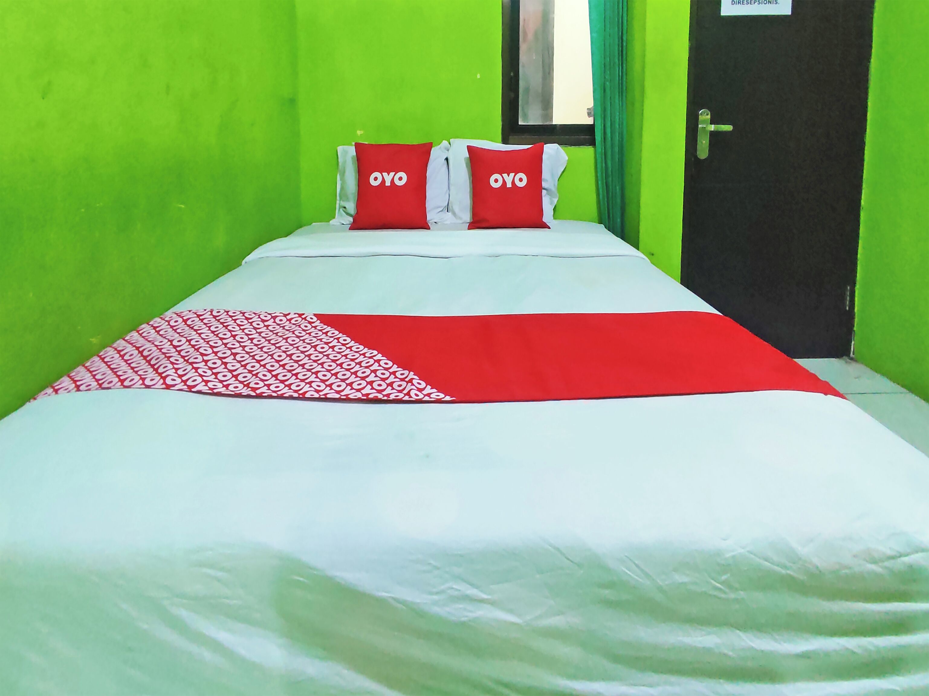 Une chambre avec un lit simple, des murs verts, une porte noire et deux oreillers sur lesquels est inscrit le mot « OYO ».