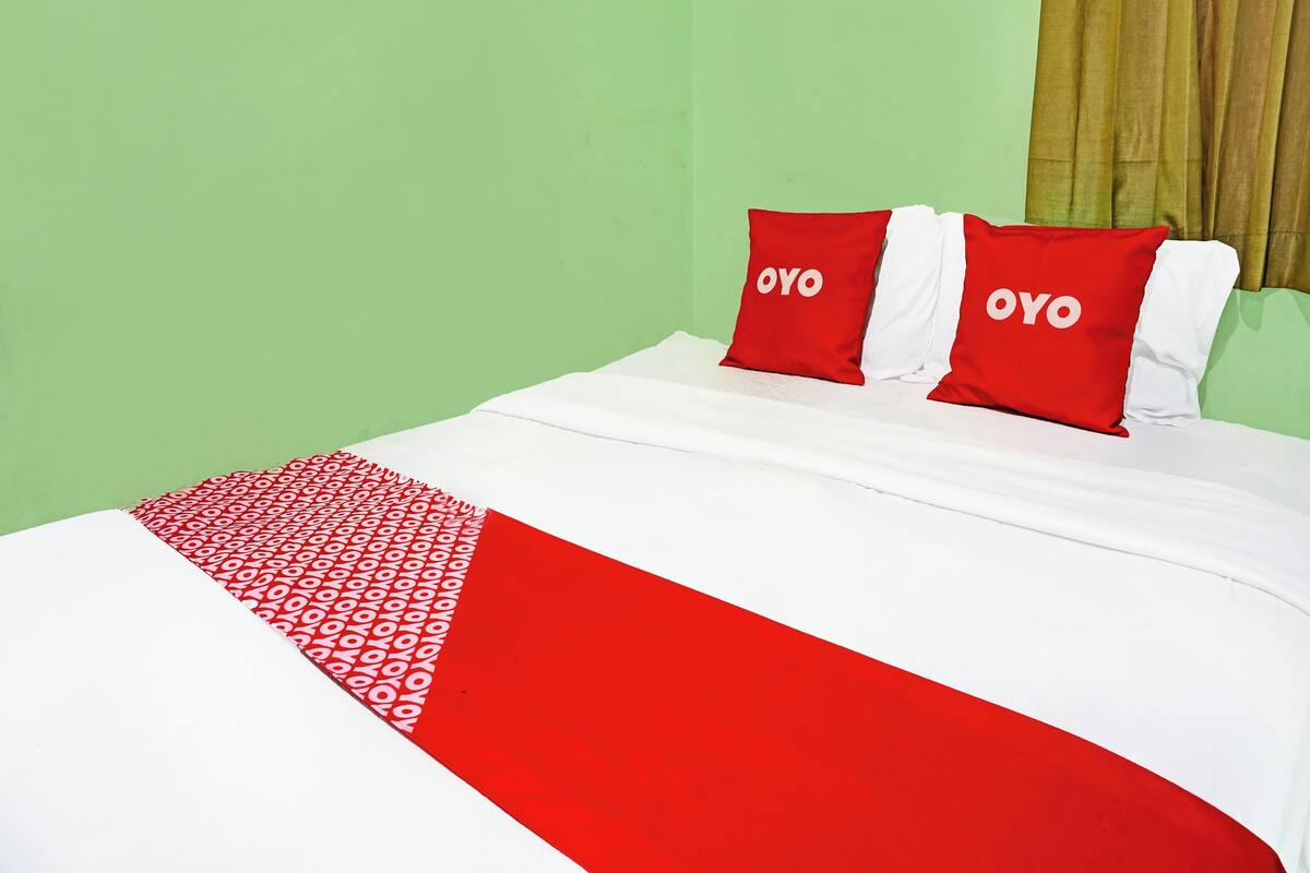 A bed with a white and red bedspread, two red pillows with the word "OYO" on them, and a green wall.