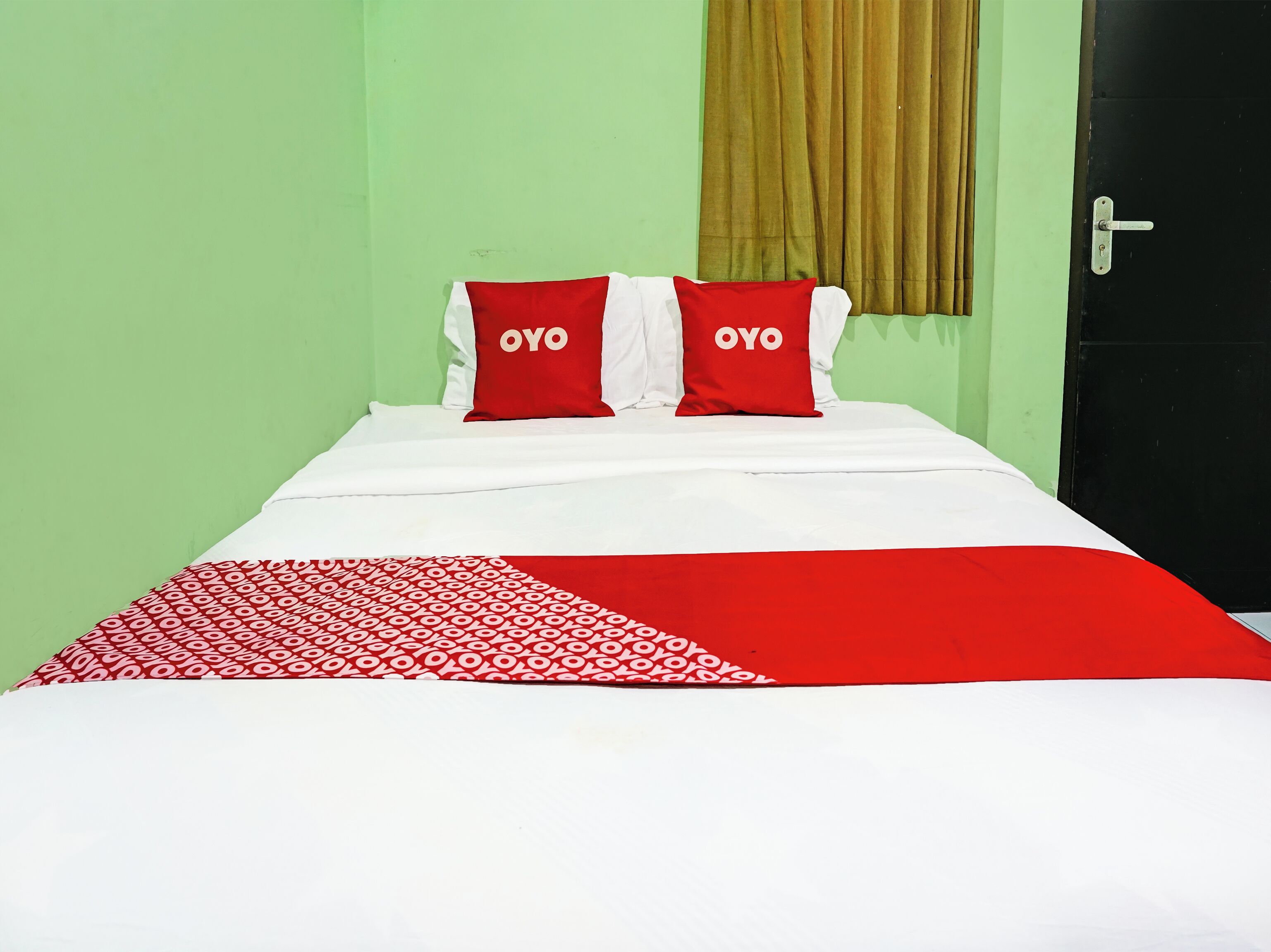 A bed with a red and white patterned blanket, two red pillows with "OYO" text, and a green wall with a brown curtain.