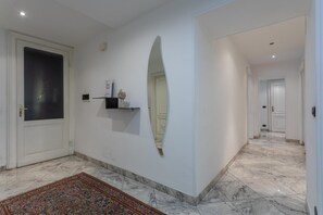 A hallway with a white door, a decorative mirror, a small shelf with items, and a patterned rug.