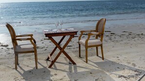Une table en bois, accompagnée de deux chaises en osier, est placée sur une plage de sable, avec l’océan en arrière-plan.