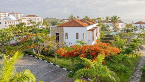 Un bâtiment blanc, surmonté d’un toit recouvert de tuiles rouges, est entouré d’une végétation luxuriante et de plantes en fleurs.