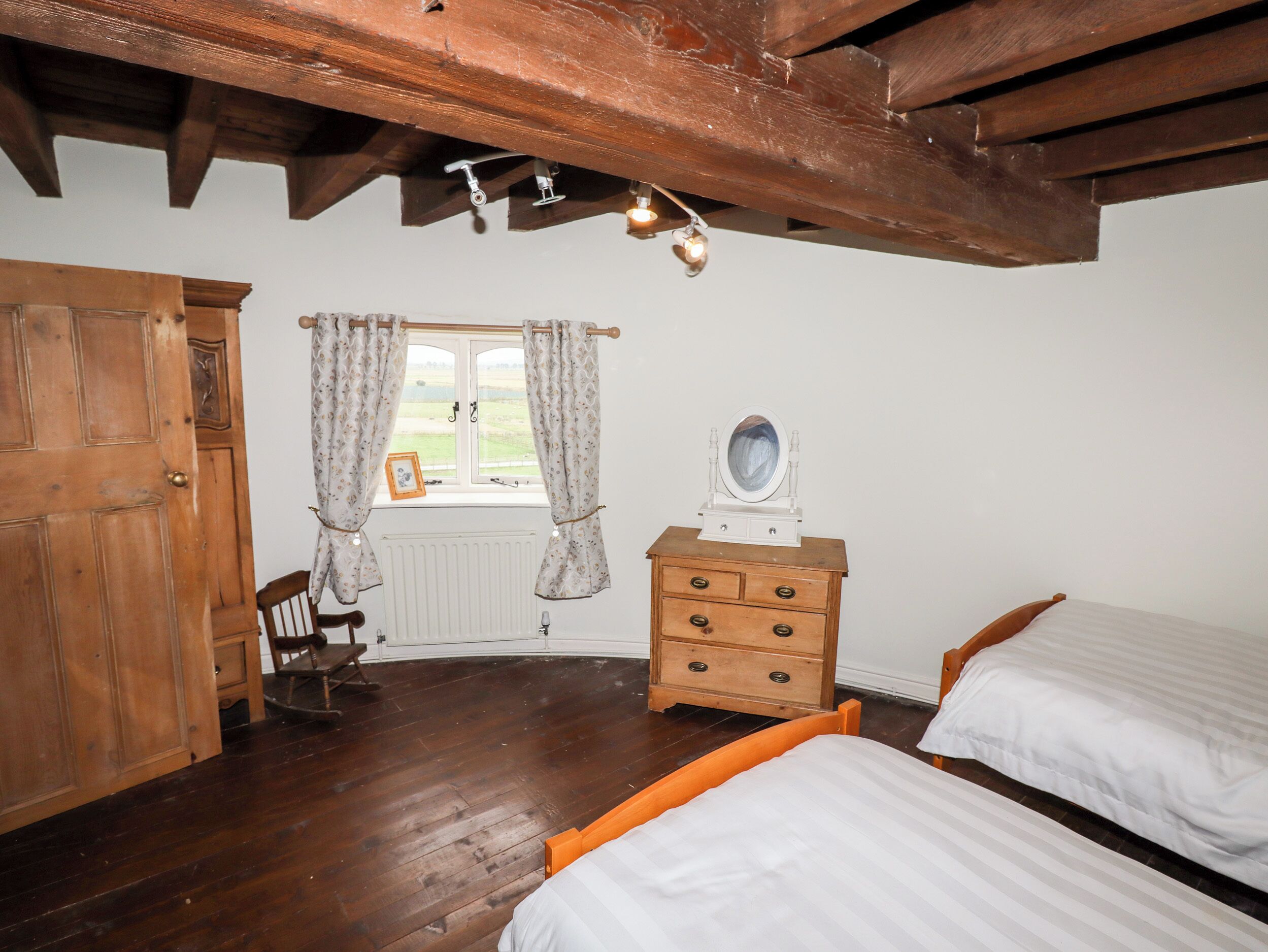 A room with wooden floors, a wooden bed, a wooden dresser with a mirror, a wooden chair, and a window with curtains.