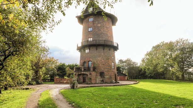 Ein Turm mit Backsteinfassade und kreisförmigem Fundament, umgeben von einer Rasenfläche und Bäumen.