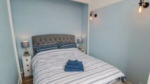 Quarto com cabeceira estofada, roupa de cama listrada, duas mesas de cabeceira com abajures e uma toalha azul sobre a cama.
