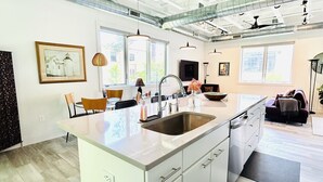 A modern kitchen with a large island, white cabinetry, and a black countertop. There are pendant lights, a window with a view, and a ceiling with exposed ductwork. - Luxury Creekside Condo , steps away from shopping & dining (Traverse City)