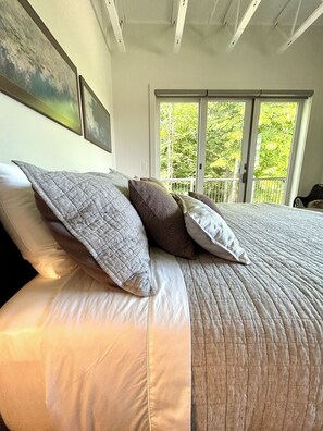 A bedroom with a large bed, a balcony with a view of trees, and a ceiling with exposed beams. - Luxury Creekside Condo , steps away from shopping & dining (Traverse City)