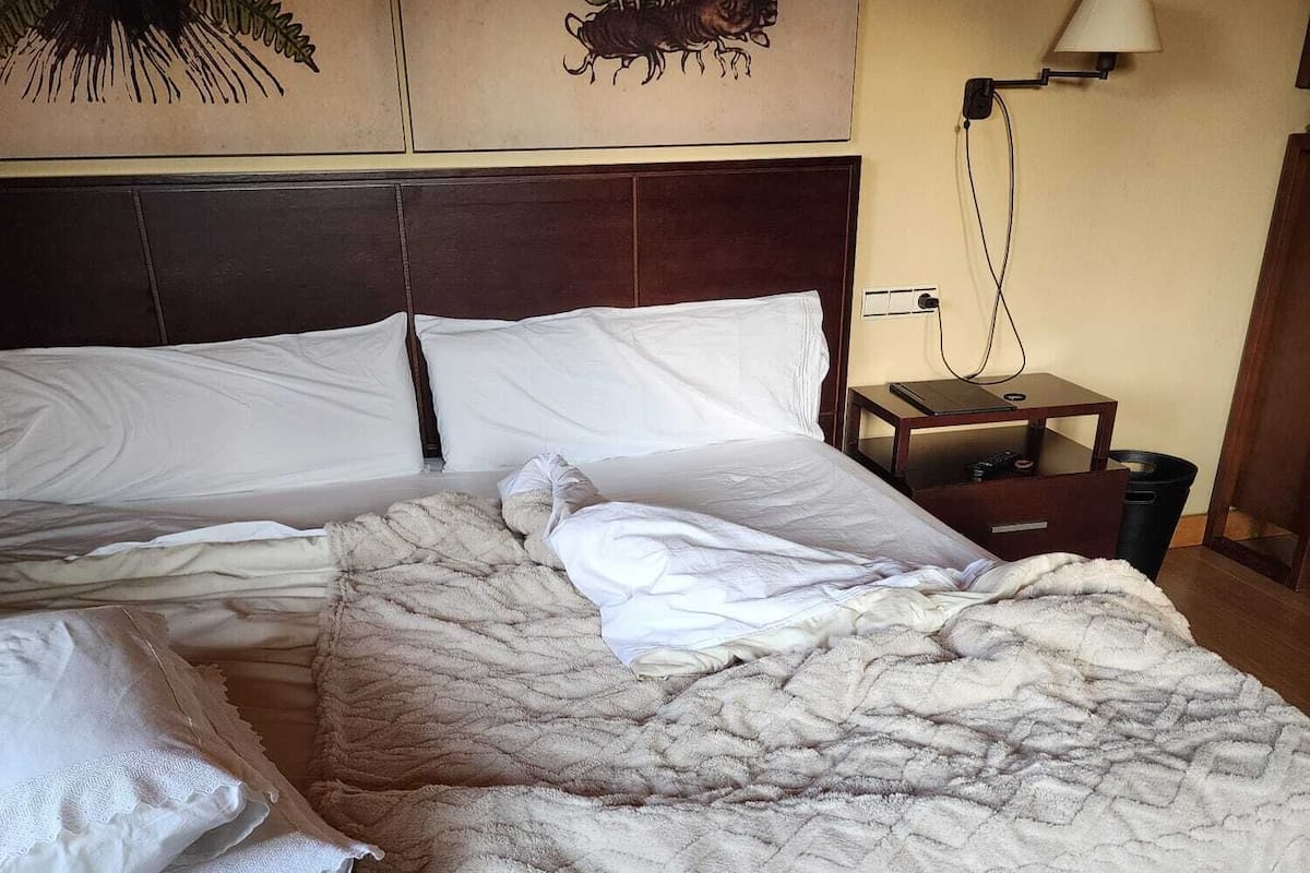 Una cama bien tendida con una colcha blanca y almohadas, un cabecero de madera, una lámpara de pared y una mesita de noche con una lámpara.