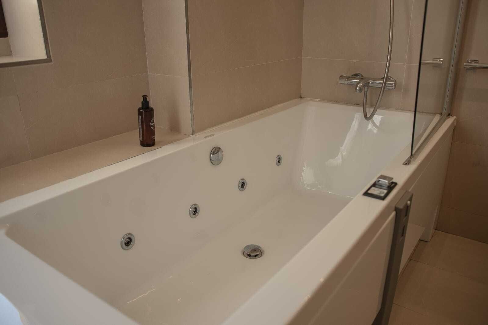 A large, rectangular bathtub with multiple faucets, a glass shower enclosure, and a small bottle of shampoo on the side.