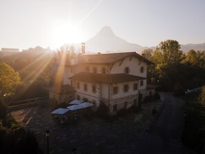Un edificio de dos pisos con tejado de tejas, rodeado de árboles y una montaña al fondo.