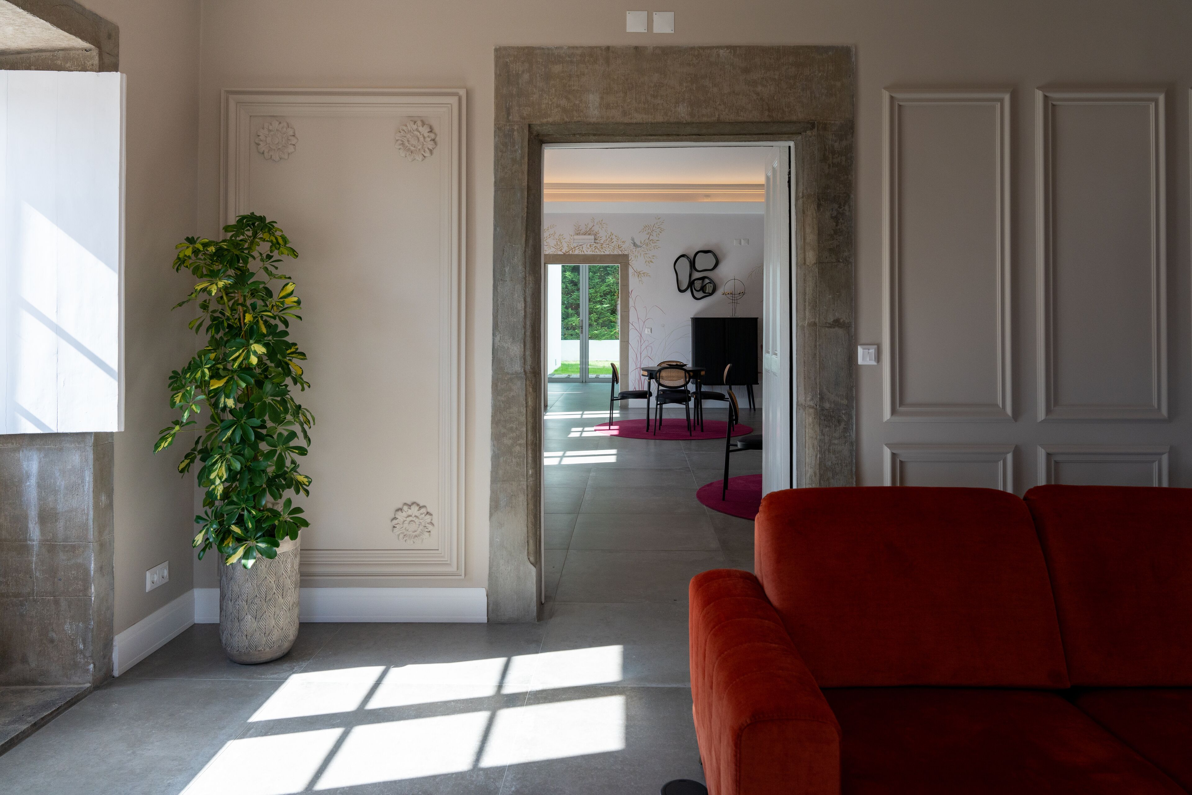 Un salon spacieux agrémenté d’une grande plante en pot, d’un canapé rouge et offrant une vue sur une autre pièce grâce à une porte ouverte.