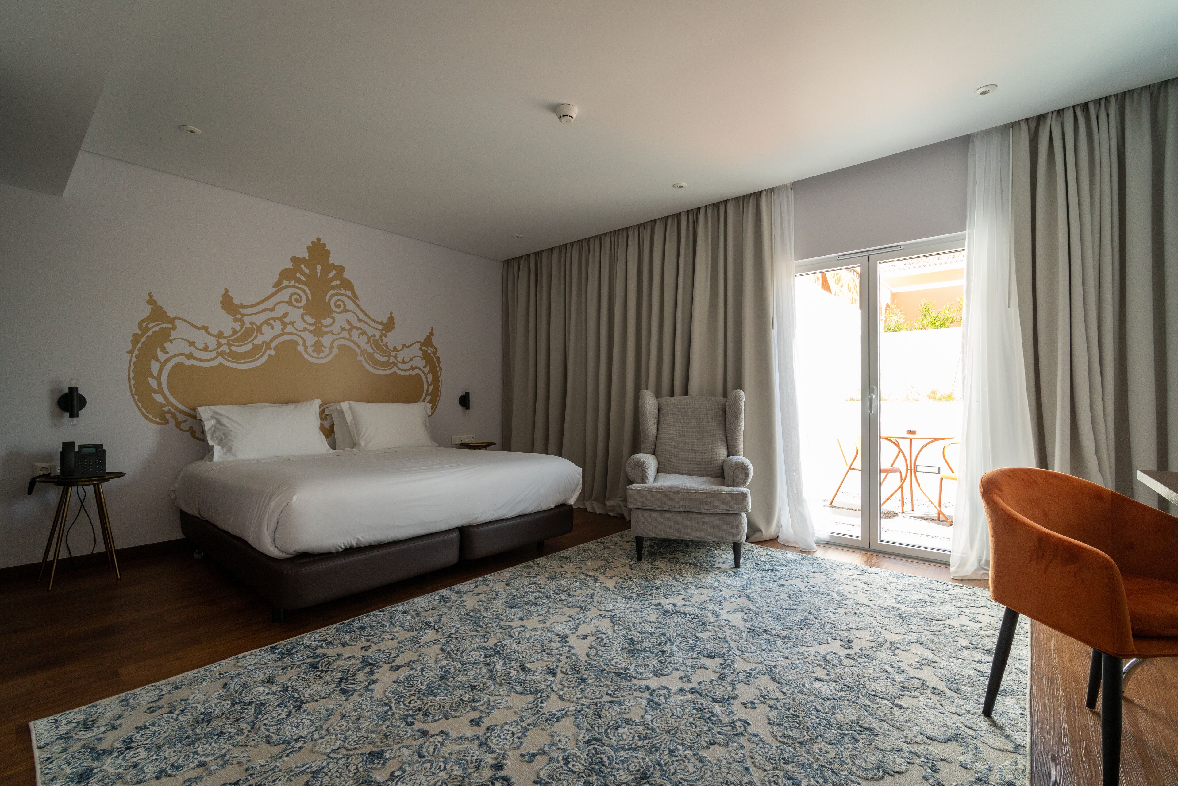 Quarto de hotel com uma cama grande, uma cadeira, uma mesinha e vista para o exterior por uma porta de correr.