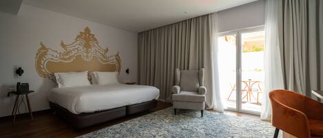 Quarto de hotel com uma cama grande, uma cadeira, uma mesinha e vista para o exterior por uma porta de correr.