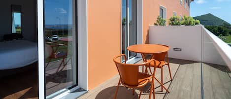 A balcony with a glass door leading to a bedroom, a small orange table and chairs, and a view of a mountain.