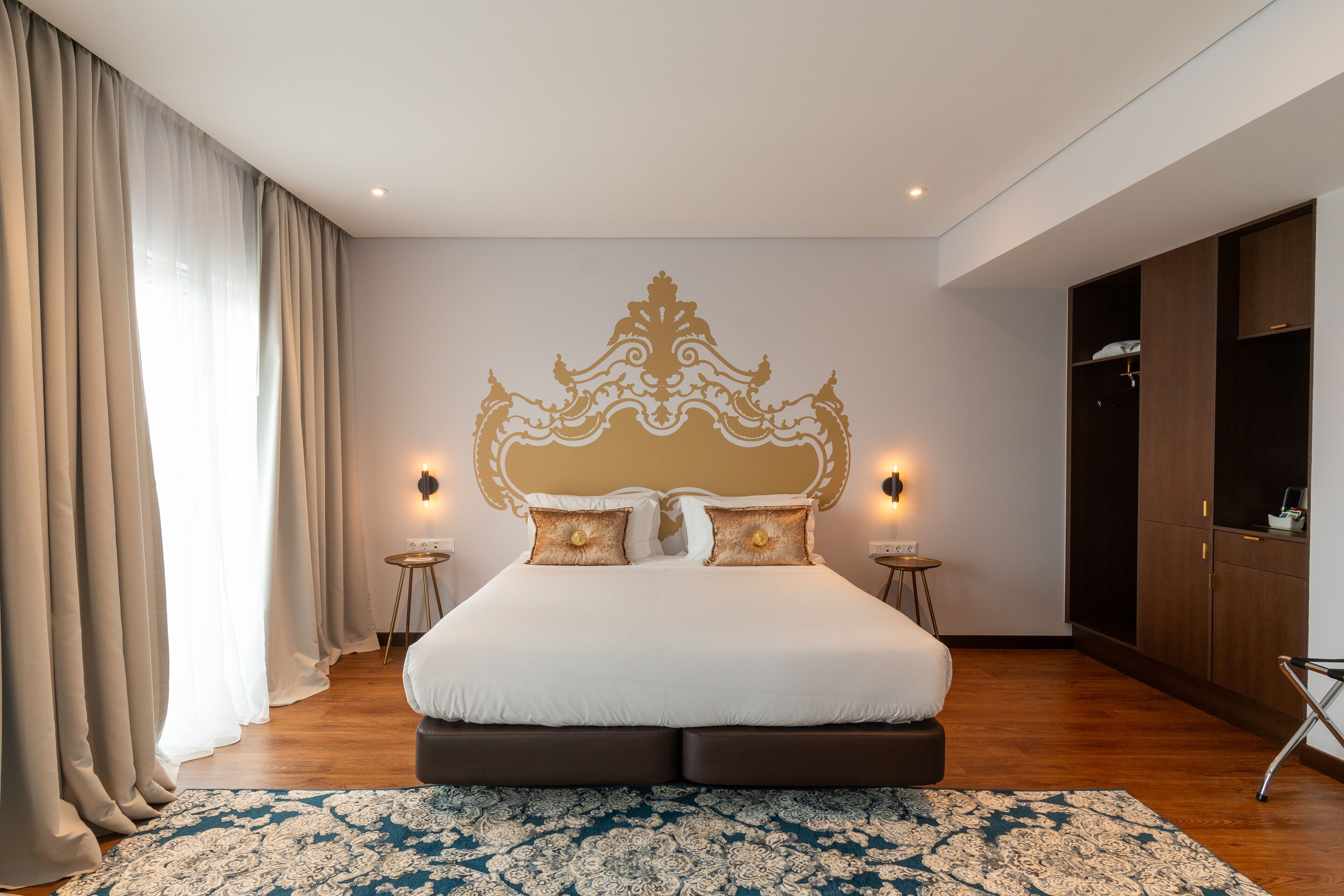 A large bed with a decorative headboard, two bedside tables, and a wardrobe in a well-lit room with wooden flooring.