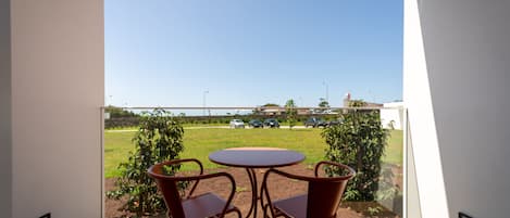A balcony with a round table and two chairs, overlooking a grassy area with trees and parked cars.