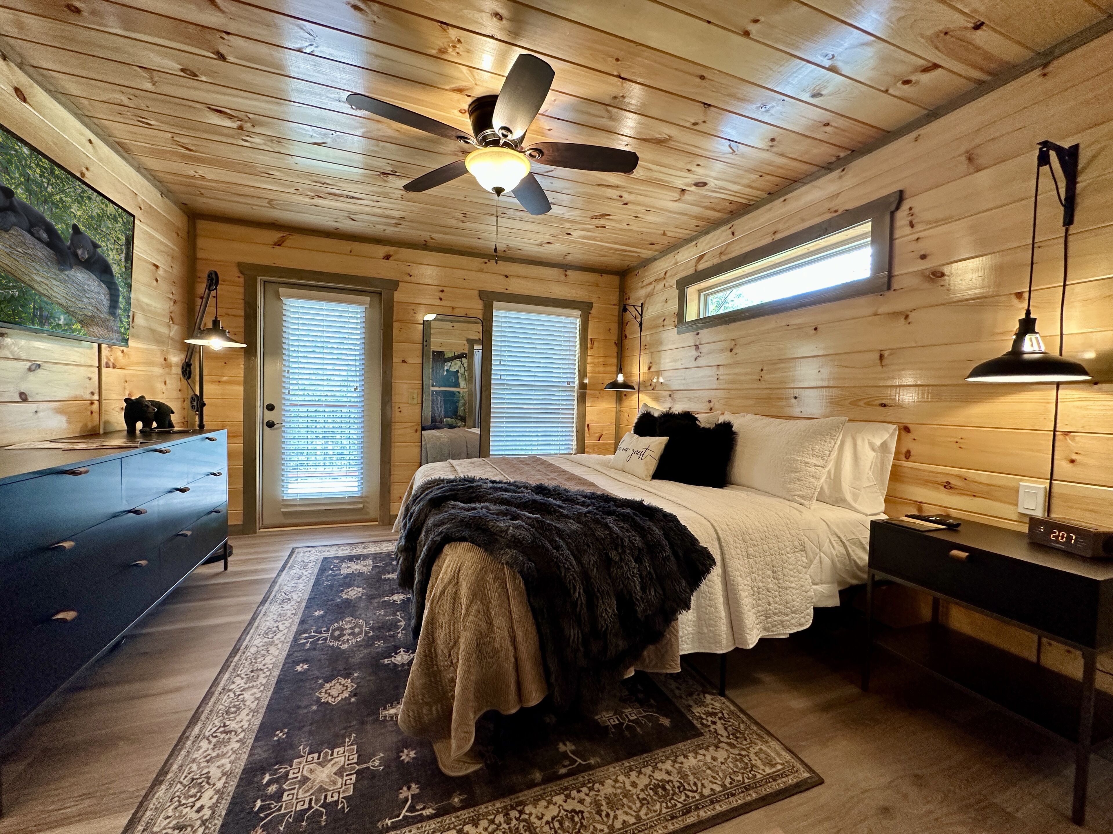 Un dormitorio de cabaña de madera con cama, cómoda, ventilador de techo y un ventanal grande.