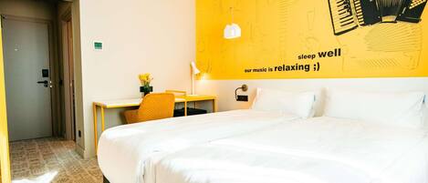 A hotel room with a large bed, a desk with a chair, a wall with musical instruments and text, and a door.