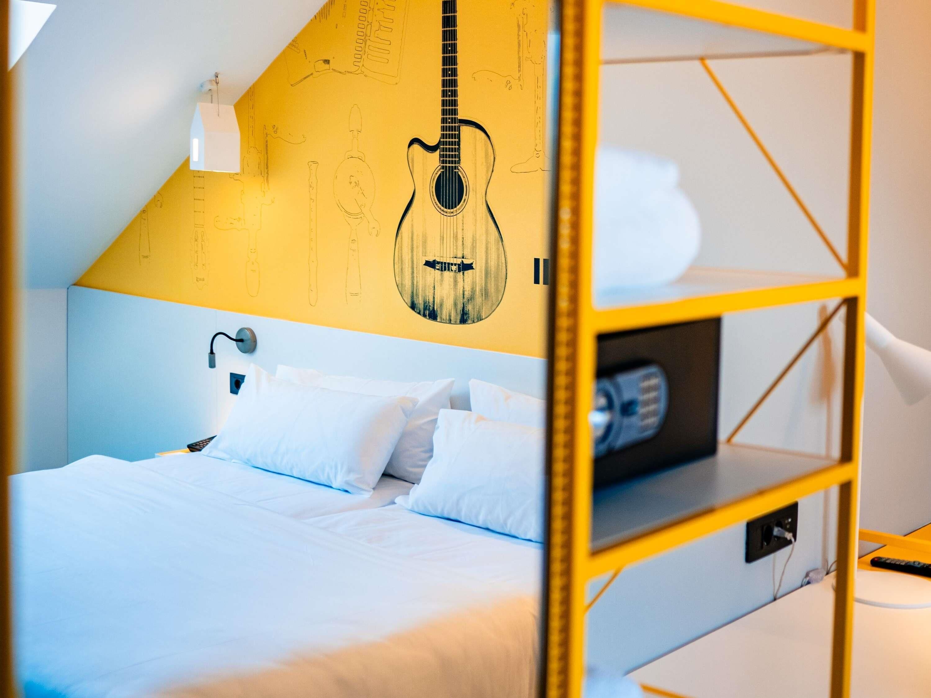 Une chambre avec un lit, des oreillers, une décoration murale représentant une guitare, une lampe et une étagère jaune.
